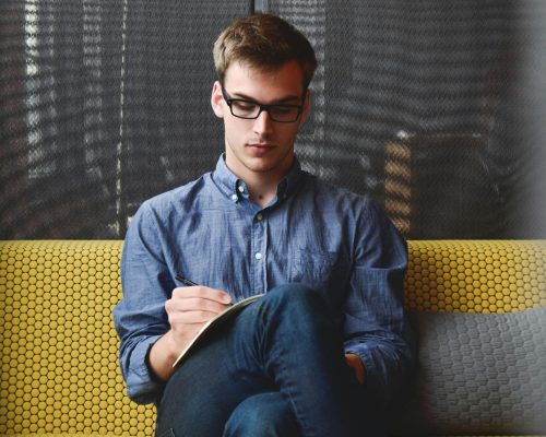 A young man in glasses writes in a notebook while sitting on a stylish couch indoors.
