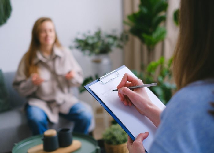 Unrecognizable professional female psychologist writing on clipboard while sitting against client on blurred background during psychotherapy session in light office